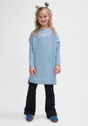 Hellblaues Sweatkleid mit langen Ärmeln und seitlichen Schlitzen, kombiniert mit schwarzen Schlaghosen und blauen Schuhen. Mit einem Logo auf der Brust.