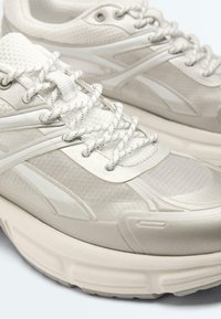 Paire de baskets athlétiques blanc cassé avec tissu en mesh texturé, lacets à motifs et semelles épaisses rembourrées.