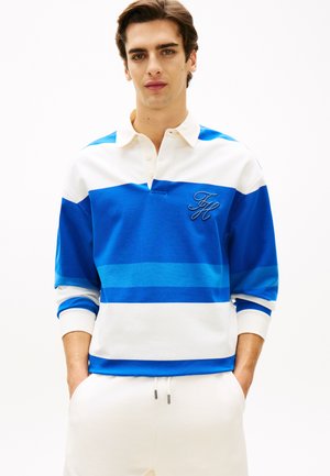 Ung mand med hænderne i lommerne iført en hvid og blåstribet rugbytrøje med broderede initialer "FH" på en ensfarvet baggrund.