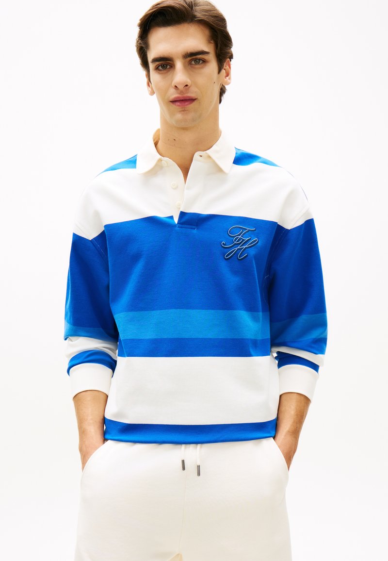 Νέος άνδρας με τα χέρια στις τσέπες, φορώντας ένα ριγέ ράγκμπι πουκάμισο σε λευκό και μπλε χρώμα με κεντημένα αρχικά "FH" σε μονόχρωμο φόντο.