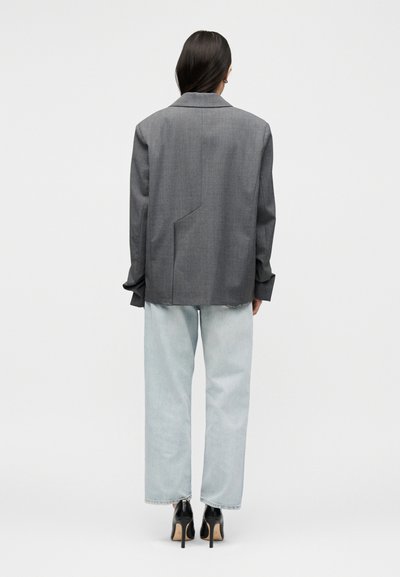 Γυναίκα με σκούρα μαλλιά που φοράει γκρι oversized σακάκι, ανοιχτό μπλε παντελόνι με φαρδιά πόδια και μαύρα ψηλοτάκουνα παπούτσια, στέκεται με την πλάτη στραμμένη.