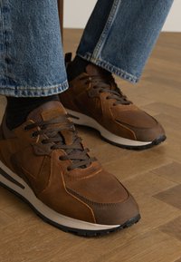 Baskets marron avec un mélange de matériaux en cuir et en maille, présentant des coutures renforcées, des lacets texturés et une semelle blanche contrastante.