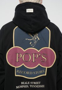 Zwarte hoodie met een grote, kleurrijke grafische afbeelding op de achterkant met de tekst "POP'S RECORD STORE" en een illustratie van een saxofoon.