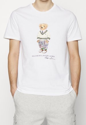 Valkoinen t-paita, jossa on karhu, joka pukeutunut neulepaitaan, ruudullisiin housuihin ja kenkiin, etupuolella teksti "POLO BEAR by RALPH LAUREN".