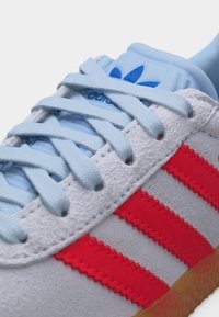 Șireturi albastru deschis pe o pantofă sport din piele întoarsă albă, cu dungi roșii în zigzag și logo-ul Adidas albastru pe limbă.