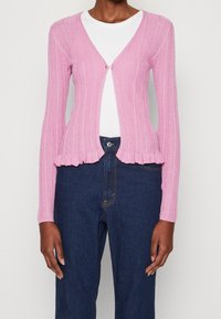 Roze geribde cardigan met een V-hals en een fraai afgewerkte onderkant, voorzien van een enkele knoopsluiting. Draagt over een witte top en in combinatie met donkere jeans.