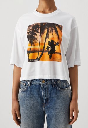 Camiseta blanca recortada con una puesta de sol naranja, palmeras y una silueta de dibujo animado, combinada con vaqueros azules.