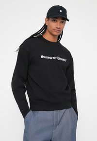Sweatshirt preto com texto branco &quot;os novos originais&quot; no peito, com decote redondo e punhos canelados. Usado com calças azuis e um boné.