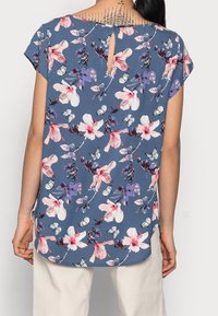 Bluzka z krótkim rękawem w niebieskim kolorze z kwiatowym wzorem, zawierająca różowe i białe kwiaty oraz motyle, z tylnym wycięciem w kształcie klucza i miękkim, teksturowanym materiałem.