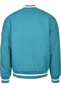 Veste universitaire bleu sarcelle avec corps en tissu uni, manches brillantes, col, poignets et ourlet côtelés ornés de rayures blanches, vue de dos.