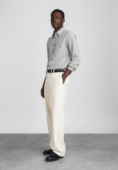 Stripet langermet skjorte med knapper i svart og hvitt, kombinert med kremfargede bukser og svarte sko, stående mot en nøytral bakgrunn.