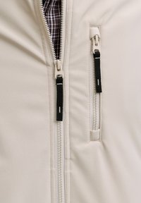 Veste beige avec une texture lisse, deux fermetures éclair noires et une doublure intérieure à carreaux. Des détails de couture remarquables améliorent son design.