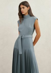 Lichtblauwe mouwloze jurk met een hoge halslijn, geplooide rok en een bijpassende riem met een rechthoekige gesp. Gladde stoftextuur.