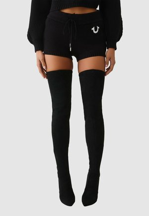 Czarne dzianinowe spodenki z prążkowanymi brzegami i ściągaczem w pasie, zestawione z wysokimi czarnymi zamszowymi kozakami sięgającymi ud. Charakterystyczne metalowe detale na spodenkach.