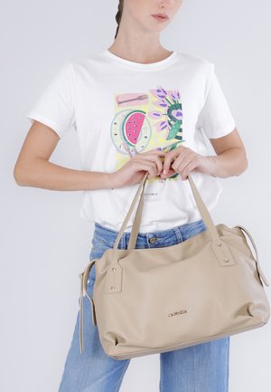 Nainen valkoisessa graafisessa t-paidassa ja sinisissä farkuissa, joka pitää suurta beigeä nahkalaukkua, jossa on kahdet kahvat.