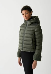 Pufferjack in donkergroen met rits aan de voorkant, capuchon en faux-fur manchetten. Beschikt over horizontale stiksels en een logo op de schouder.