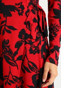 Röd klänning i tyg med svart blommönster och knytbälte, med fokus på midjan och en hand som vilar vid kjolen.