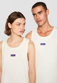 Dos personas llevan camisetas sin mangas de color crema claro con un logo "GANT" en azul y rojo en el pecho; textura suave y acanalada.