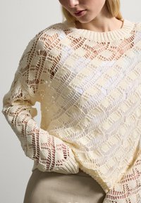Jersey de punto en un suave y cremoso amarillo con un patrón geométrico de calado y cuello acanalado. Tiene mangas largas con acentos texturizados.