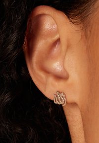 Zilveren studoorbel met een gestileerd ontwerp in een gladde, gepolijste afwerking, geplaatst tegen een close-up van een oor met een gestructureerde huid.