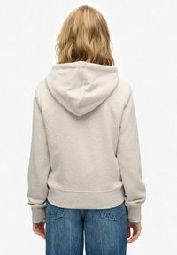 Sudadera beige con capucha ajustable, textura suave y puños y dobladillo acanalados, combinada con vaqueros azules de corte holgado, vista desde la parte de atrás.