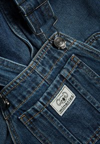 Denim tuinbroek in donkerblauw met koperen stiksel, voorzien van verstelbare banden en een ronde metalen sluiting. Het label toont een koala-afbeelding.