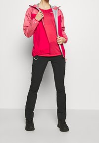 Punainen ja vaaleanpunainen kevyt takki, jossa on harmaa huppu, yhdistetty mustiin ulkohousuihin ja mustiin saappaisiin, suunniteltu ulkoiluun.