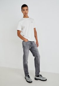 Λευκό βαμβακερό T-shirt με μικρό λογότυπο, συνδυασμένο με γκρι τζιν παντελόνι και παπούτσια γκρι με μοτίβο. Καθαρές γραμμές και άνετη εφαρμογή.