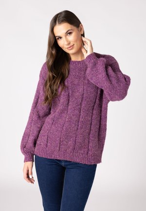 Maglione lavorato a maglia viola con un motivo a treccia e maniche ampie. Indossato con jeans scuri su uno sfondo neutro.