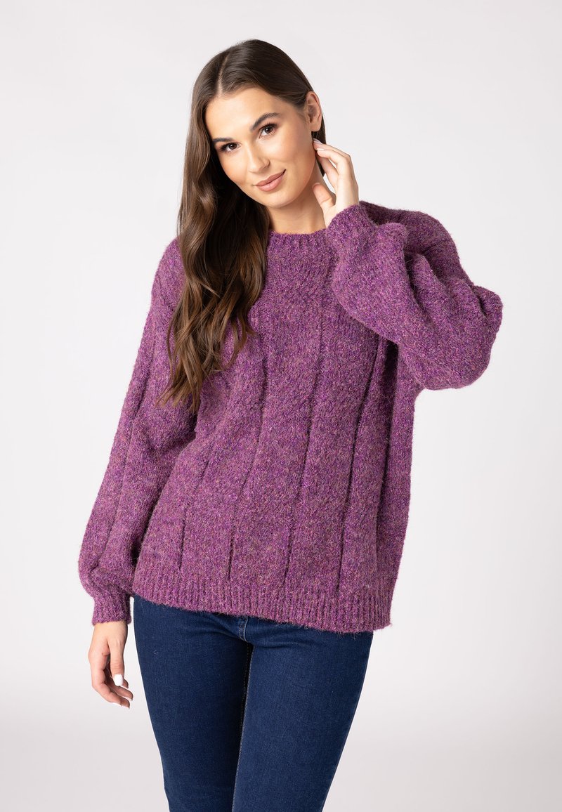Lila gestrickter Pullover mit einem strukturierten Zopfmuster und weiten Ärmeln. Wird mit dunklen Jeans vor einem schlichten Hintergrund getragen.