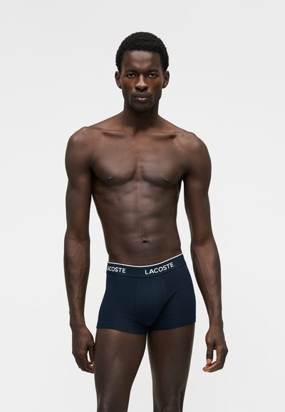 Фит тъмнокож мъж с къса къдрава коса, облечен в тъмносини боксерки Lacoste, който стои пред светъл едноцветен фон.