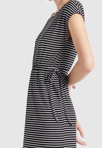 Kvinnas svart och vit horisontellt randig klänning med korta ärmar och ett knytet midjebälte, visad från sidan mot en enkel bakgrund.