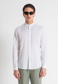 Camicia bianca a maniche lunghe con colletto alla coreana, abbinata a occhiali da sole neri. Tessuto in lino liscio, vestibilità su misura e colore chiaro.
