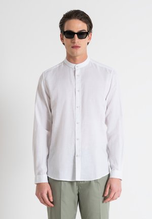 Camicia bianca a maniche lunghe con colletto alla coreana, abbinata a occhiali da sole neri. Tessuto in lino liscio, vestibilità su misura e colore chiaro.