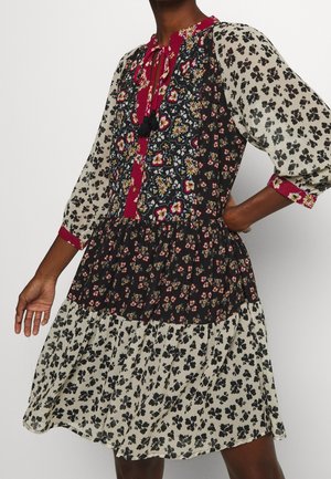 Femme portant une robe à motifs floraux multiples avec des accents rouges, des manches trois-quarts et une taille froncée, debout devant un fond uni.