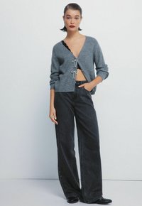 Szary dzianinowy kardigan z głębokim dekoltem w kształcie V i wiązaniem, w połączeniu z czarnymi dżinsami o wysokim stanie i luźnym kroju, z bocznymi guzikami.