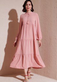 Robe rose à manches longues et superposées avec un col montant et des détails à franges. Tissu léger et silhouette fluide. Accentuée par des sandales colorées.