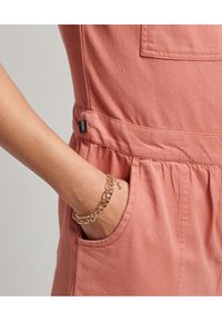 Różowy bawełniany kombinezon z boczną kieszenią i subtelną metką marki. Złota bransoletka łańcuszkowa zdobi nadgarstek, podkreślając kontrast materiałów.
