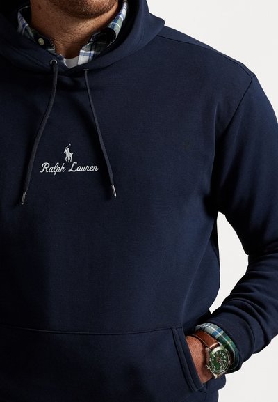 Uomo che indossa un hoodie navy di Ralph Lauren sopra una camicia a quadri verde, con una mano in tasca e un orologio con cinturino in pelle marrone ben visibile.