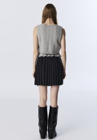 Femme debout, de dos, portant un haut sans manches en tricot gris à mailles, une jupe plissée noire et des bottes noires montantes.