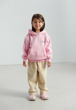 Roza pamučna hoodica s prednjim džepom i plavim tekstom, uparena s bež trenirkom i svijetloružastim klompkama s rupama. Jednostavan i ležeran outfit.