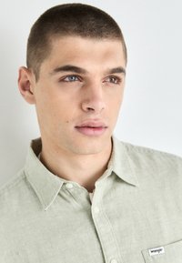 Light green, button-up shirt made of textured fabric with a collar and a pocket featuring a small "Wrangler" label.