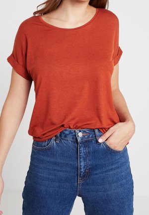 Kvinde iført en rustfarvet kortærmet skjorte stoppet ned i højttaljede blå denimjeans med den ene hånd i lommen.