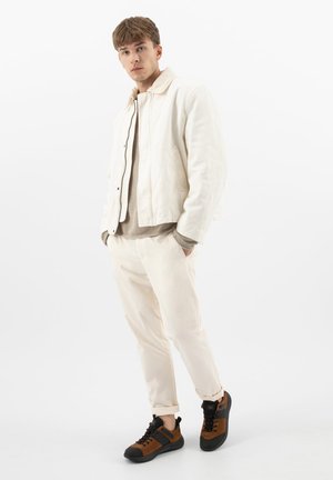 Młody mężczyzna stojący z rękami w kieszeniach, ubrany w beżową kurtkę, jasne spodnie, brązowo-czarne trampki na jednolitym białym tle.