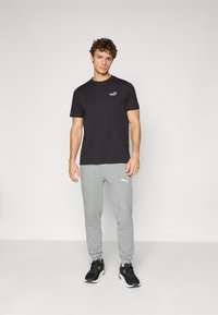 Zwarte katoenen T-shirt met een klein goudkleurig logo, gecombineerd met lichtgrijze joggers met een wit logo, en zwarte sportschoenen met groene accenten.