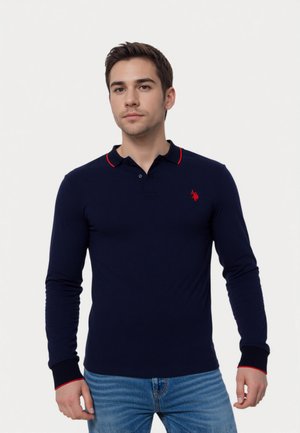 Jonge man draagt een marineblauwe lange mouwen polo met rode bies aan de kraag en manchetten, gecombineerd met lichtblauwe jeans, staande tegen een witte achtergrond.
