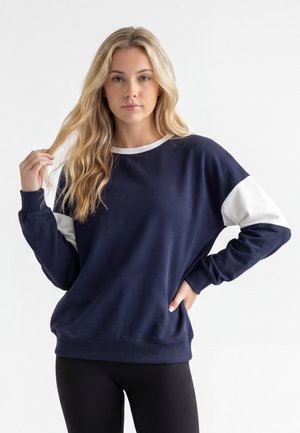 Femme blonde portant un sweat-shirt bleu marine avec des panneaux blancs aux manches, debout avec une main dans la poche et l'autre touchant ses cheveux.