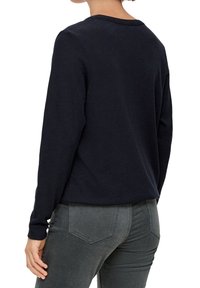 Marinblå långärmad t-shirt med rundad halsringning. Tyget verkar mjukt; figurnära design med rak rygg. Bärs tillsammans med gråa byxor.