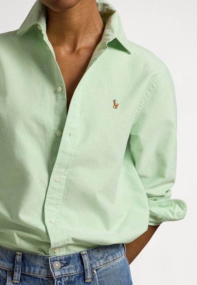 Gaiši zaļa kokvilnas krekls ar pogām, klasiska apkakle un maza izšūta zirga logo uz krūtīm, valkāts kopā ar zilām džinsa biksēm.