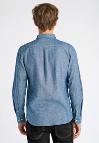 Heren overhemd met lange mouwen van lichtblauwe stof, met een button-down kraag, een enkele plooi op de rug en knoopmanchetten, gecombineerd met donkere jeans.